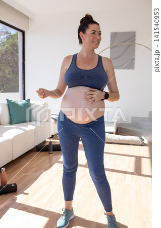 Pregnant woman placing hand on belly during living room workout with yoga mat and water bottles Pregnant woman placing hand on belly during living room workout with yoga mat and water bottles 141540953