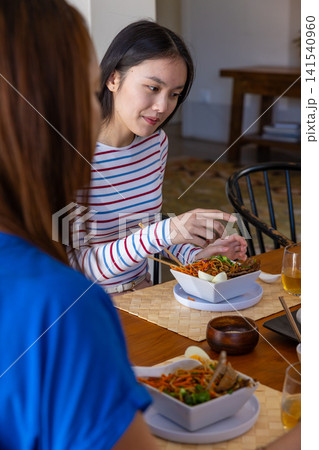 Diverse female friends sitting at dining table eating noodle salad with chopsticks, amber beverage 141540960