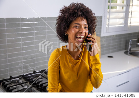 Woman leaning on counter laughing, talking on smartphone near gas stovetop 141541009