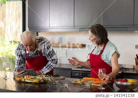 Senior couple cooking at kitchen island wearing red aprons using tablet while chopping vegetables Senior couple cooking at kitchen island wearing red aprons using tablet while chopping vegetables 141541138