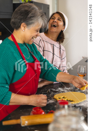 Senior mother and daughter rolling dough with red cutters at kitchen counter near wine glass Senior mother and daughter rolling dough with red cutters at kitchen counter near wine glass 141541148