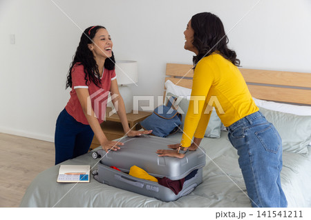Diverse mom and teenage daughter packing silver suitcase in bedroom with notebook, pen, backpack Diverse mom and teenage daughter packing silver suitcase in bedroom with notebook, pen, backpack 141541211