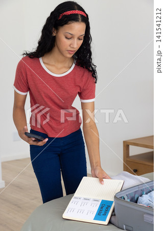Teenage girl checking packing checklist, holding smartphone next to open suitcase in bedroom 141541212