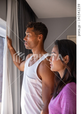 Diverse couple at window in bedroom, pulling white curtain beside drape and peering outside Diverse couple at window in bedroom, pulling white curtain beside drape and peering outside 141541239