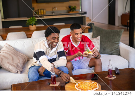 Diverse male friends smiling and grabbing pizza from board with soda bottles in living room Diverse male friends smiling and grabbing pizza from board with soda bottles in living room 141541274