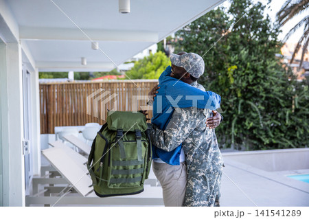 African american father and son embracing on patio by table with camo uniform, green backpack African american father and son embracing on patio by table with camo uniform, green backpack 141541289