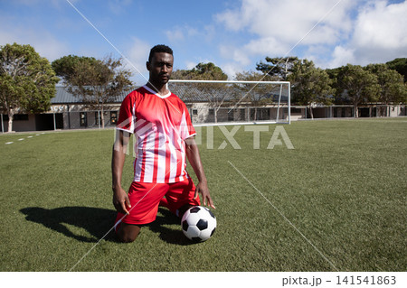 African American male soccer player kneeling on field by goalpost resting on ball in red jersey 141541863