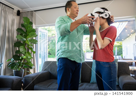 Asian father with daughter helping put on white virtual reality headset in living room 141541900