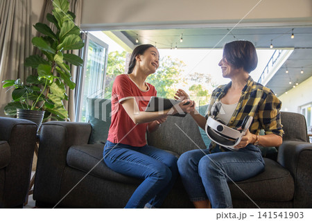 Hispanic mother with asian daughter laughing while using tablet and vr headset at home 141541903