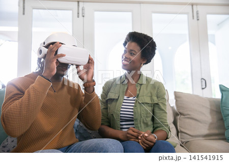 African american couple sitting on couch at home, adjusting white vr headset while woman smiling African american couple sitting on couch at home, adjusting white vr headset while woman smiling 141541915