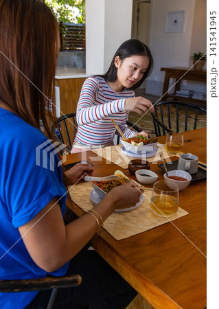 Asian mother and daughter at kitchen table eating from bowls with chopsticks and iced tea 141541945