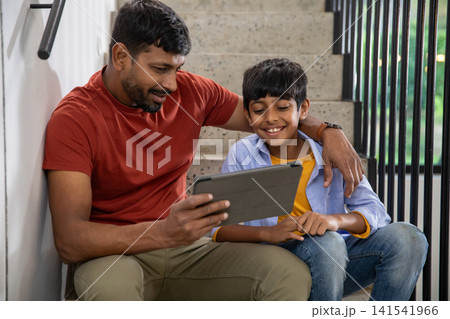 Indian father and son sitting on staircase landing at home and viewing tablet computer together 141541966