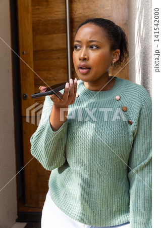 African american woman wearing sweater at entryway holding smartphone near mouth, speaking African american woman wearing sweater at entryway holding smartphone near mouth, speaking 141542000
