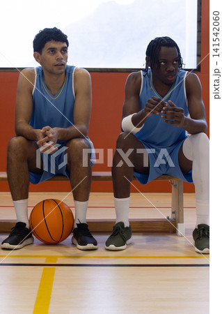 Diverse teammates sitting on bench in gym in light blue uniforms and gesturing near basketball Diverse teammates sitting on bench in gym in light blue uniforms and gesturing near basketball 141542060