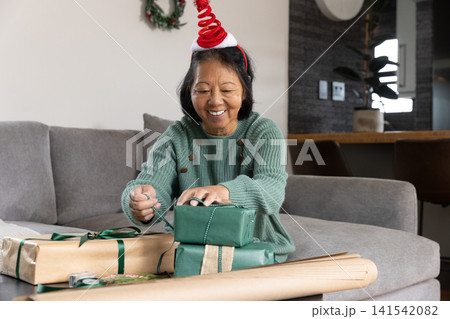 Senior asian woman in red headband wrapping gifts on coffee table in living room with wreath Senior asian woman in red headband wrapping gifts on coffee table in living room with wreath 141542082
