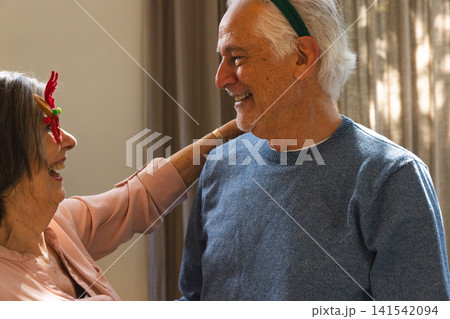 Senior couple standing face to face in warm living room wearing red novelty glasses, green headband Senior couple standing face to face in warm living room wearing red novelty glasses, green headband 141542094