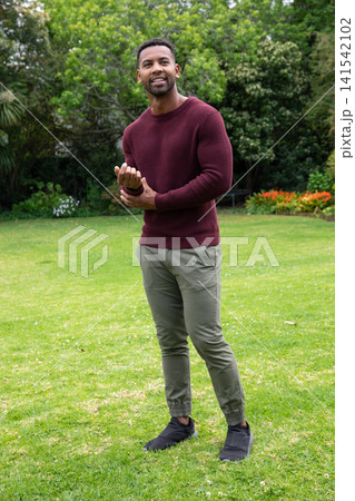 African american man standing on garden lawn adjusting burgundy sweater cuff beside blooms African american man standing on garden lawn adjusting burgundy sweater cuff beside blooms 141542102