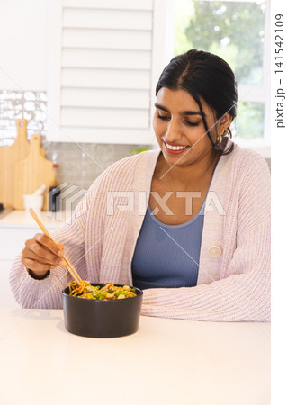 South Asian woman eating vegetable noodles from black bowl with wooden chopsticks at kitchen island 141542109