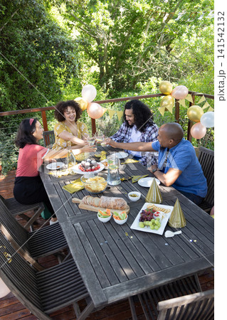 Diverse friends raising wine glasses at backyard deck table with pastel balloons, banner, cake Diverse friends raising wine glasses at backyard deck table with pastel balloons, banner, cake 141542132