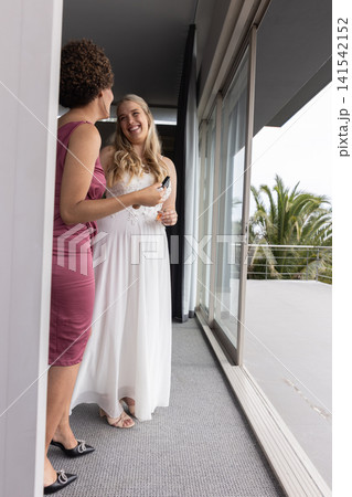 Diverse bride and bridesmaid chatting at sliding door, with smartphone boutonniere by railing 141542152