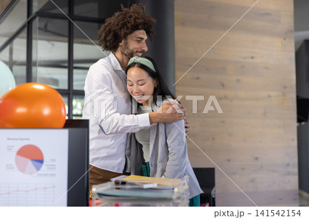 Diverse coworkers hugging beside office desk in casual wear with monitor showing charts, balloon Diverse coworkers hugging beside office desk in casual wear with monitor showing charts, balloon 141542154
