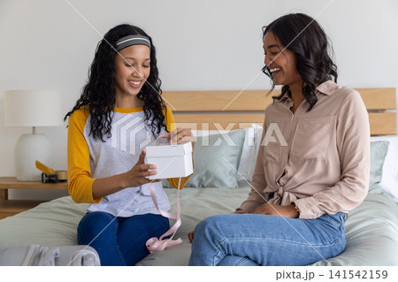 Diverse mom and teenage daughter sitting on bed at home untying pink ribbon gift box smiling Diverse mom and teenage daughter sitting on bed at home untying pink ribbon gift box smiling 141542159