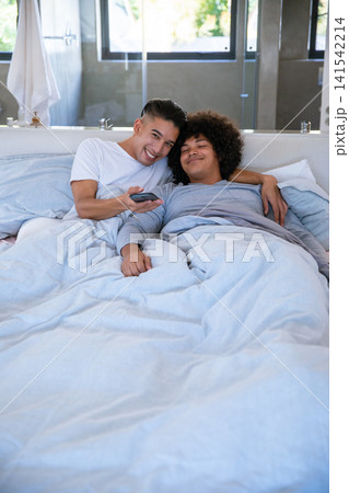 Two men reclining on bed at home holding TV remote control under duvet with pillows 141542214