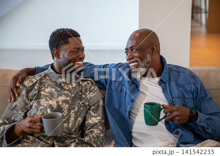 African american father and son smiling on couch at home with mugs, son wearing camouflage 141542235