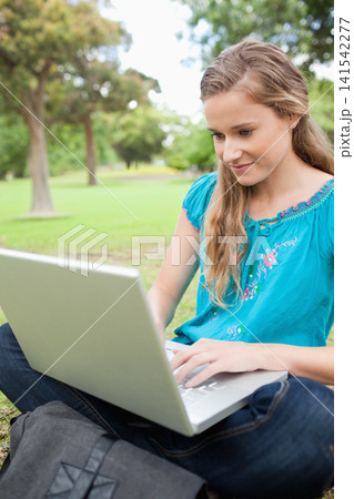 Female wearing teal top and jeans, sitting cross-legged on park grass using silver laptop, backpack Female wearing teal top and jeans, sitting cross-legged on park grass using silver laptop, backpack 141542277