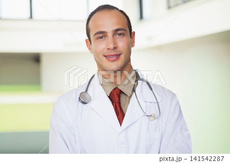 Male physician standing in medical facility lobby wearing white lab coat red tie stethoscope Male physician standing in medical facility lobby wearing white lab coat red tie stethoscope 141542287