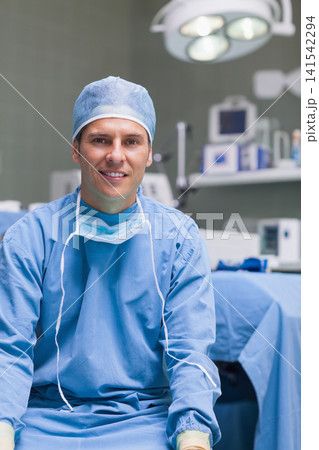 Mid-adult male surgeon smiling, sitting in operating room in blue scrubs and cap, mask around neck Mid-adult male surgeon smiling, sitting in operating room in blue scrubs and cap, mask around neck 141542294