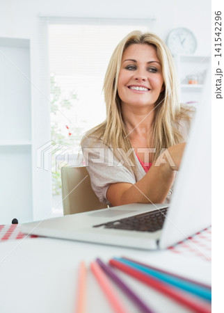 Woman smiling while working on laptop at table at home with checkered cloth and pencils Woman smiling while working on laptop at table at home with checkered cloth and pencils 141542296