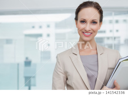 Woman standing right holding binder in beige blazer facing camera in office by windows, copy space Woman standing right holding binder in beige blazer facing camera in office by windows, copy space 141542336
