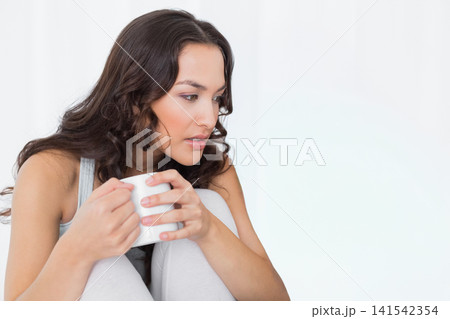 Woman in twenties sitting left in lit room wearing light loungewear holding white ceramic mug 141542354