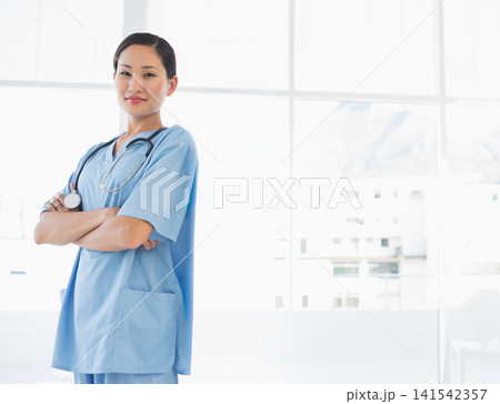 Asian woman standing with arms crossed at clinic wearing light blue scrubs, stethoscope, copy space Asian woman standing with arms crossed at clinic wearing light blue scrubs, stethoscope, copy space 141542357