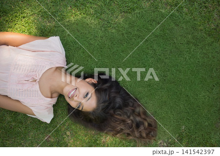 Asian adult woman lying on green lawn smiling, spreading long wavy hair, wearing pink blouse Asian adult woman lying on green lawn smiling, spreading long wavy hair, wearing pink blouse 141542397