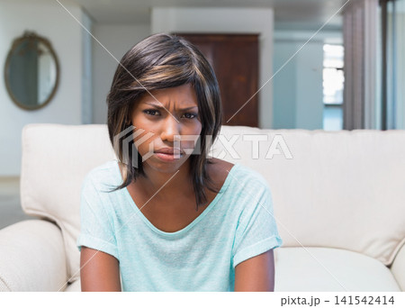 African American woman sitting on beige sofa in living room, frowning and holding cushion African American woman sitting on beige sofa in living room, frowning and holding cushion 141542414