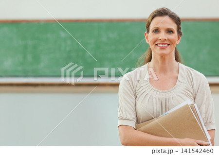 Woman teacher holding papers, tan folder by green board classroom front with chalk tray, copy space Woman teacher holding papers, tan folder by green board classroom front with chalk tray, copy space 141542460