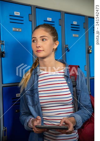 Teenage girl wearing denim jacket using tablet in school hall by lockers with backpack 141542473