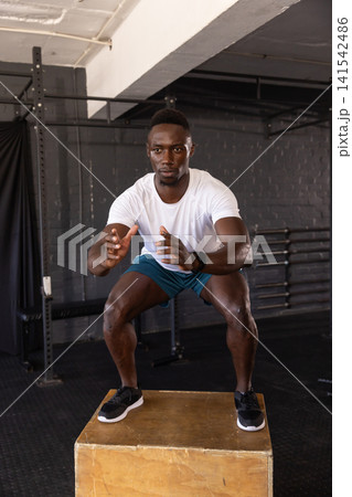 African American man doing plyometric box jump at gym with wooden box, squat rack and smartwatch 141542486