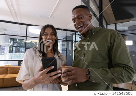 Diverse coworkers laughing and sharing smartphone while holding coffee mug in office lounge 141542509