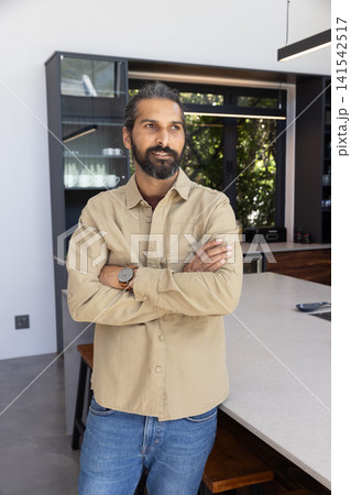 Mid adult indian man leaning on kitchen island countertop in kitchen, holding remote Mid adult indian man leaning on kitchen island countertop in kitchen, holding remote 141542517