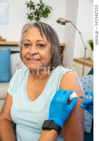 Senior hispanic woman sitting on couch getting vaccination from gloved nurse with syringe, pad Senior hispanic woman sitting on couch getting vaccination from gloved nurse with syringe, pad 141542533