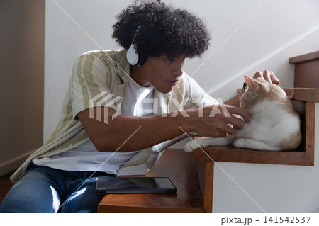 African american man on stairs with white headphones petting cat beside tablet at home African american man on stairs with white headphones petting cat beside tablet at home 141542537