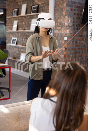 Diverse female coworkers wearing white vr headset and raising hands in office lounge Diverse female coworkers wearing white vr headset and raising hands in office lounge 141542580