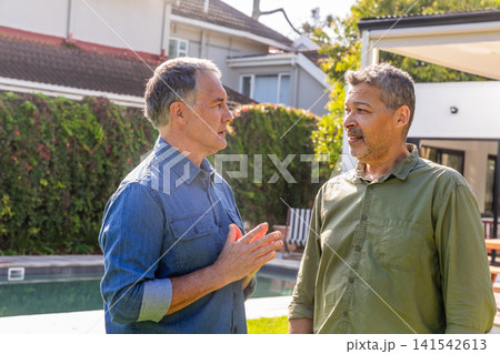 Two male friends standing by in-ground pool in suburban backyard, chatting near patio furniture Two male friends standing by in-ground pool in suburban backyard, chatting near patio furniture 141542613