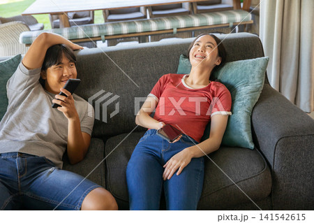 Asian friends reclining on sofa with teal cushion at home using smartphones and smiling 141542615