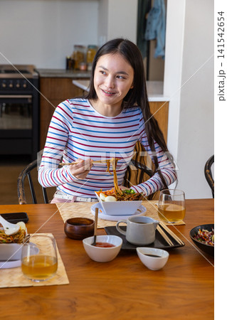 Asian woman eating noodles from bowl with chopsticks at table in kitchen with smartphone Asian woman eating noodles from bowl with chopsticks at table in kitchen with smartphone 141542654