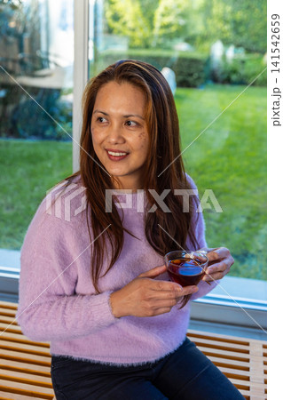 Mature asian woman wearing lavender sweater holding glass tea cup, sitting by large window Mature asian woman wearing lavender sweater holding glass tea cup, sitting by large window 141542659