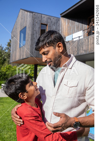 South asian father in wristwatch holding son on green lawn before wooden house with laundry 141542668